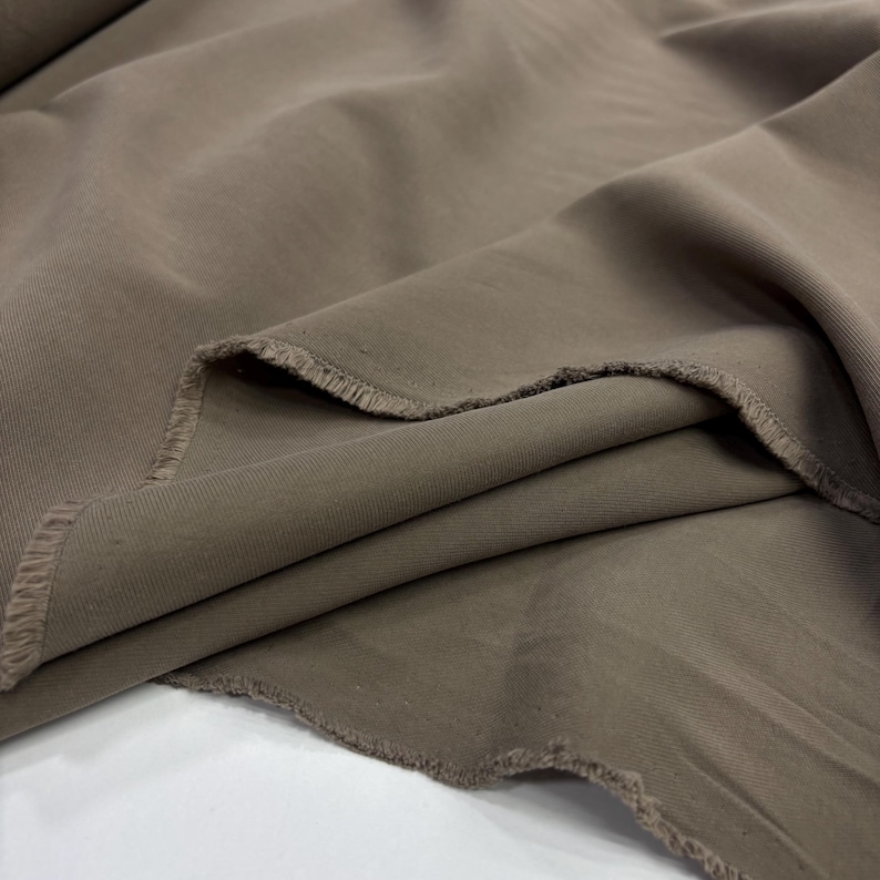 May include: A length of solid taupe-colored fabric with a visible diagonal twill weave pattern. The material is draped in soft folds, revealing a raw, frayed edge along the cut section of the textile, showcasing its opaque and matte texture.