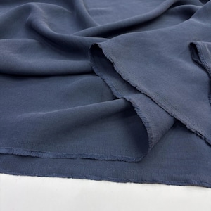 May include: A length of solid dark blue fabric with a visible weave texture, featuring soft folds and drapes. The edges of the material are raw and slightly frayed, showing the unfinished cut of the textile as it rests in loose, flowing layers.