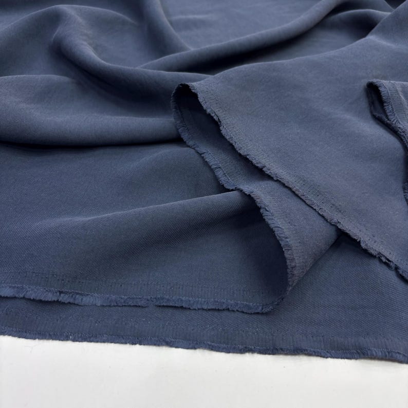 May include: A length of solid dark blue fabric with a visible weave texture, featuring soft folds and drapes. The edges of the material are raw and slightly frayed, showing the unfinished cut of the textile as it rests in loose, flowing layers.