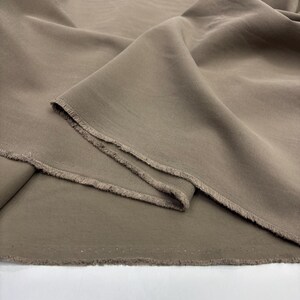 May include: A length of solid taupe-colored fabric featuring a visible twill weave texture. The material is draped in soft folds, showcasing a raw, unfinished edge with loose threads along the cut side of the textile piece.