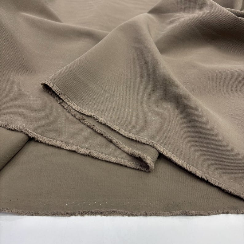 May include: A length of solid taupe-colored fabric featuring a visible twill weave texture. The material is draped in soft folds, showcasing a raw, unfinished edge with loose threads along the cut side of the textile piece.