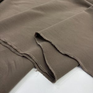 May include: A length of solid taupe-colored fabric featuring a visible diagonal twill weave pattern. The material is folded to show its drape, with raw, unfinished edges displaying loose threads along the cut side of the textile piece.
