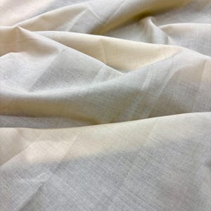 May include: Close-up of a light-colored fabric with subtle texture and folds. The fabric appears to be a blend of off-white and pale gray, creating a soft, neutral palette. The material has a slightly translucent quality.