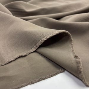 May include: A length of taupe-colored fabric with a visible twill weave pattern. The material features soft, flowing folds and drapes, showcasing a matte finish and raw, slightly frayed edges along the cut sections of the textile material.