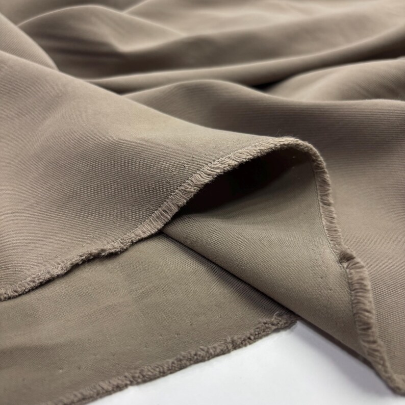 May include: A length of taupe-colored fabric with a visible twill weave pattern. The material features soft, flowing folds and drapes, showcasing a matte finish and raw, slightly frayed edges along the cut sections of the textile material.