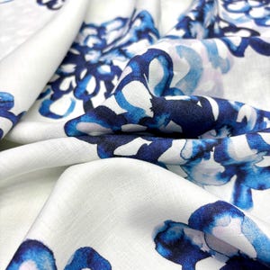 May include: Close-up of white fabric with a blue floral pattern. The design features watercolor-style blooms in shades of blue, creating a soft, artistic look. The fabric is folded, showing the texture and depth of the material.