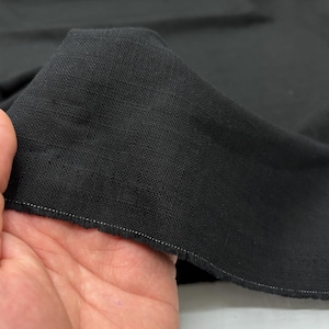 Può includere: Primo piano di tessuto nero, tenuto per mostrare la trama e l'ordito. Il tessuto ha un bordo grezzo con una linea di cucitura bianca. Il materiale appare morbido e si drappeggia bene, suggerendo che potrebbe essere utilizzato per abbigliamento o arredamento.