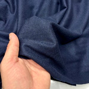May include: Close-up of a navy blue fabric, with visible texture and folds. The fabric is held in a hand, showcasing its drape and color. The material appears soft and possibly suitable for apparel or home decor.