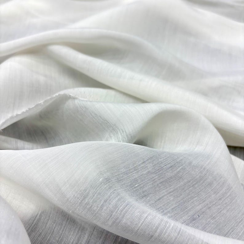May include: Close-up of white fabric with a soft, flowing texture. The material is draped in gentle folds, creating shadows and highlights. The fabric appears lightweight and slightly translucent, suggesting a delicate weave.