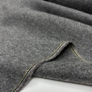 May include: Close-up of a folded piece of gray fabric. The fabric has a soft, textured appearance. The edge of the fabric is visible, showing a detailed seam with yellow and red stitching. The overall look is of a high-quality textile.