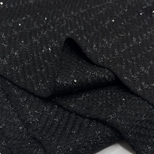 Puede incluir: Primer plano de tela negra con tejido texturizado y lentejuelas plateadas dispersas. La tela está doblada, creando sombras y resaltando la textura. El material parece ser un textil lujoso, adecuado para ropa de noche u ocasiones especiales.