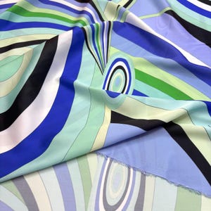 Op de afbeelding: Een levendige stof met een kleurrijk abstract ontwerp. Het patroon bevat gebogen lijnen en vormen in tinten blauw, groen, zwart en wit. De stof is gevouwen en toont de textuur en de vloeiing van het materiaal.