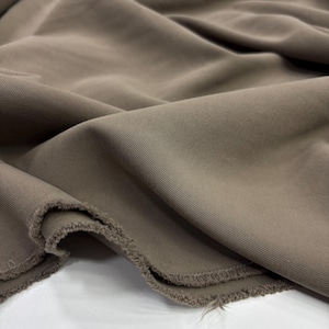 May include: A length of solid taupe-colored fabric featuring a distinct diagonal twill weave pattern. The material is draped in soft folds, showcasing the smooth texture of the surface and the raw, unfinished edges along the bottom hem.
