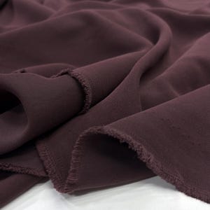 May include: A length of solid dark plum colored fabric with a visible twill weave texture. The material is draped in soft folds, showcasing the matte finish and the raw, slightly frayed edges along the cut sections of the textile material.