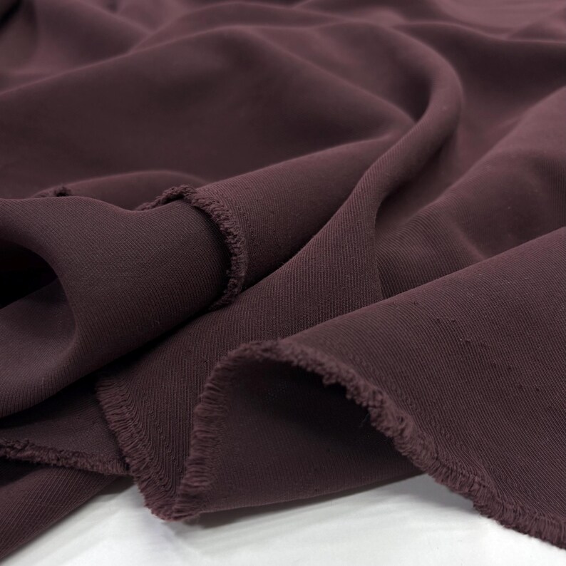 May include: A length of solid dark plum colored fabric with a visible twill weave texture. The material is draped in soft folds, showcasing the matte finish and the raw, slightly frayed edges along the cut sections of the textile material.