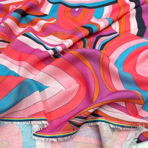 May include: A colourful fabric with an abstract design. The pattern features swirling lines and shapes in shades of pink, orange, blue, and black. The fabric has a soft, flowing appearance, with a fringed edge.