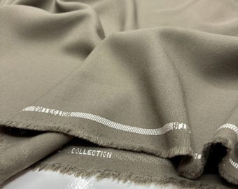 May include: Close-up of a folded, taupe-colored fabric with a textured weave. The fabric has a raw edge with the word "COLLECTION" printed in white. White stripes run along the edge of the fabric.