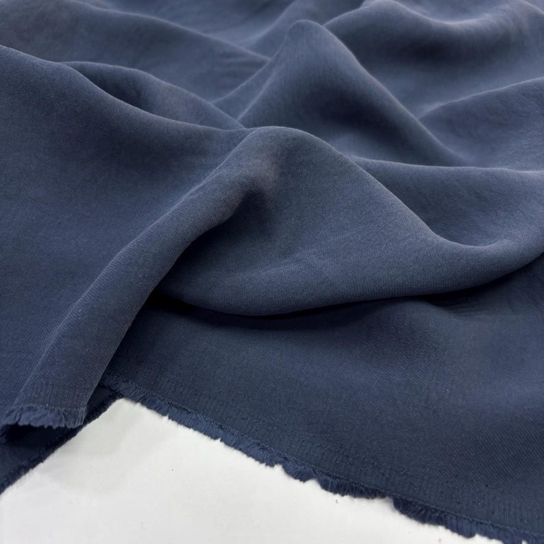 May include: A length of dark navy blue fabric with a soft, matte texture and a slightly translucent quality. The material is draped in loose, flowing folds, revealing a raw, frayed edge along the bottom border of the textile piece.