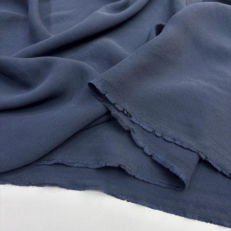 May include: A length of solid slate blue fabric with a soft, matte texture and visible folds. The material features raw, frayed edges along the cut sections, showcasing a loose weave and a consistent, muted blue color throughout the textile.