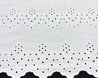 May include: White eyelet fabric with a scalloped edge. The fabric features a repeating pattern of floral and geometric cutouts, creating an openwork design. The eyelets are accented with small, evenly spaced black dots.
