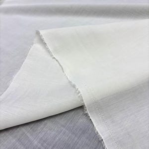 May include: Close-up of white fabric with a slightly textured surface. The fabric is folded, creating a layered effect. The edges are unfinished, showing a frayed texture. The fabric appears lightweight and suitable for textile projects.