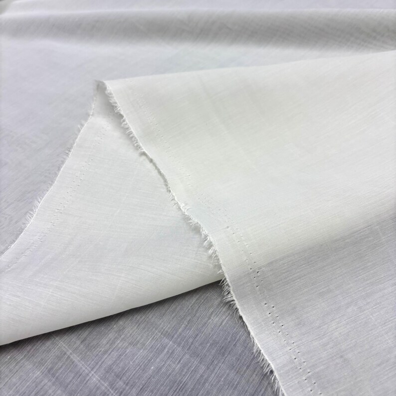 May include: Close-up of white fabric with a slightly textured surface. The fabric is folded, creating a layered effect. The edges are unfinished, showing a frayed texture. The fabric appears lightweight and suitable for textile projects.