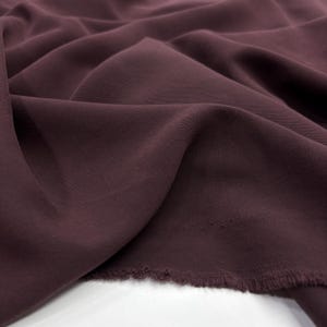May include: A deep plum-colored fabric with a visible diagonal twill weave texture. The material is draped in soft, flowing folds, creating natural shadows and highlights across the surface. The bottom edge shows a raw, unfinished frayed hem.