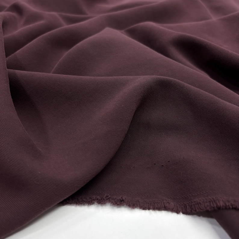 May include: A deep plum-colored fabric with a visible diagonal twill weave texture. The material is draped in soft, flowing folds, creating natural shadows and highlights across the surface. The bottom edge shows a raw, unfinished frayed hem.