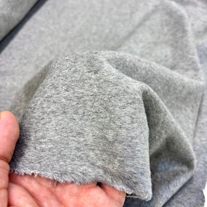 Puede incluir: Primer plano de una tela gris con una superficie suave y texturizada. La tela se sostiene, mostrando sus pliegues y el borde sin rematar. El material parece ser un textil de peso medio, adecuado para varios proyectos de costura.