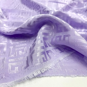 May include: Close-up of a lavender fabric with a geometric pattern. The fabric is draped, showing folds and texture. The edges are frayed, indicating a cut piece of material.