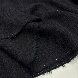 Può includere: Primo piano di un tessuto nero strutturato con un motivo intrecciato. Il tessuto ha un bordo sfilacciato con fili bianchi. Il materiale sembra spesso e adatto per progetti di abbigliamento o decorazione della casa.