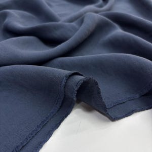 May include: A length of solid dark slate blue fabric with a matte finish and a visible woven texture. The material is draped in soft, flowing folds, showcasing the depth of the color and the slightly frayed, raw edges along the cut side of the textile.
