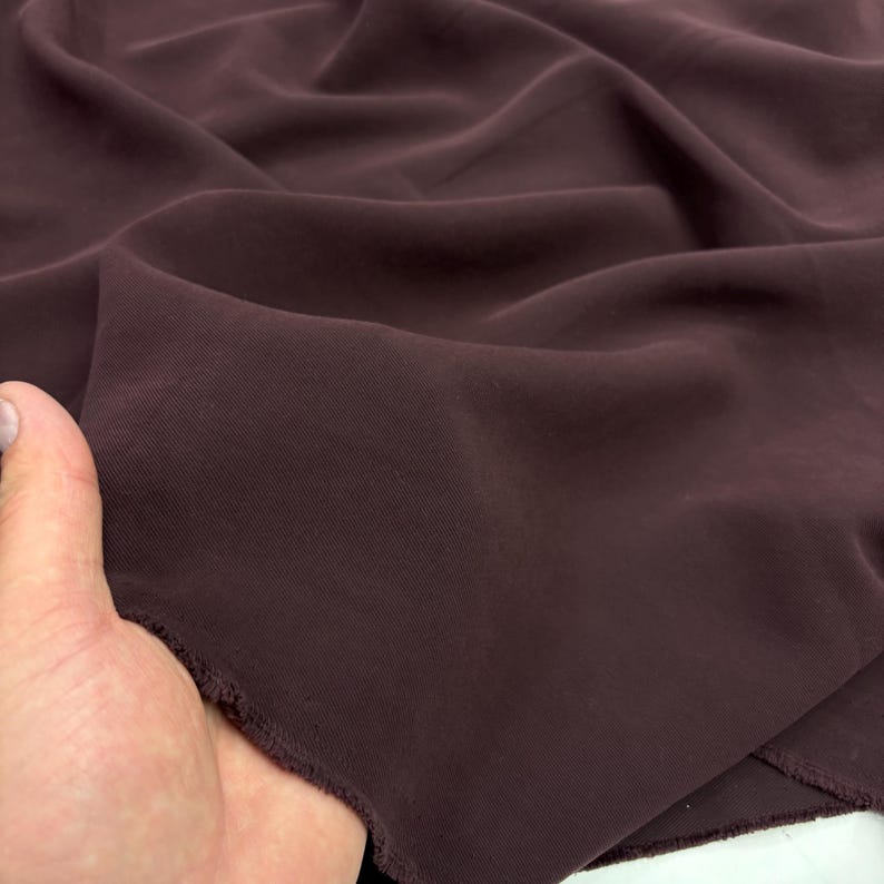 May include: A piece of solid dark plum colored fabric with a smooth, matte texture. The material is draped in soft folds, showing a consistent deep purple hue throughout the surface. The edges of the textile appear raw and unfinished.