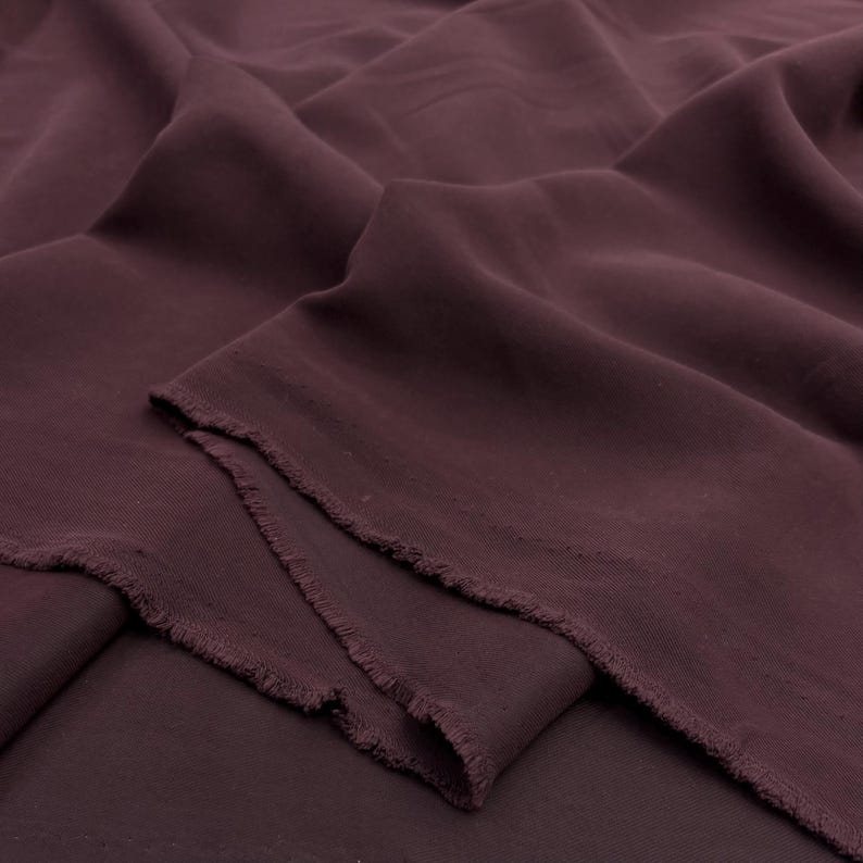 May include: A length of solid dark plum colored fabric with a smooth, matte texture. The material is draped in soft, flowing folds, creating deep shadows and highlights across the surface. The edges of the fabric show a raw, frayed cut finish.