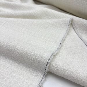 May include: A roll of off-white fabric featuring a textured, woven surface with a visible cross-hatch pattern. The material has a slightly nubby, irregular appearance with a frayed edge showing loose threads along the side of the textile.