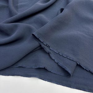 May include: A length of solid slate blue fabric with a soft, matte texture and visible folds. The material features raw, unfinished edges with a slight fringe along the cut sides, showcasing the weave and drape of the textile in a natural light.