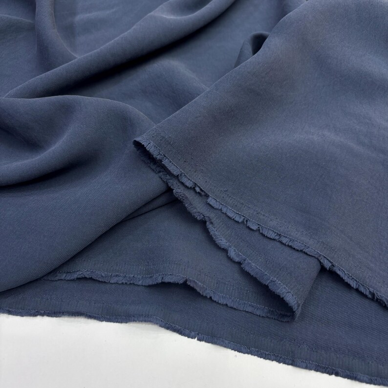 May include: A length of solid slate blue fabric with a soft, matte texture and visible folds. The material features raw, unfinished edges with a slight fringe along the cut sides, showcasing the weave and drape of the textile in a natural light.