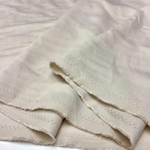May include: A length of off-white, unbleached cotton fabric featuring a visible twill weave texture. The material is folded in soft, cascading layers, revealing a raw, frayed selvage edge with a series of small, evenly spaced pinhole marks.