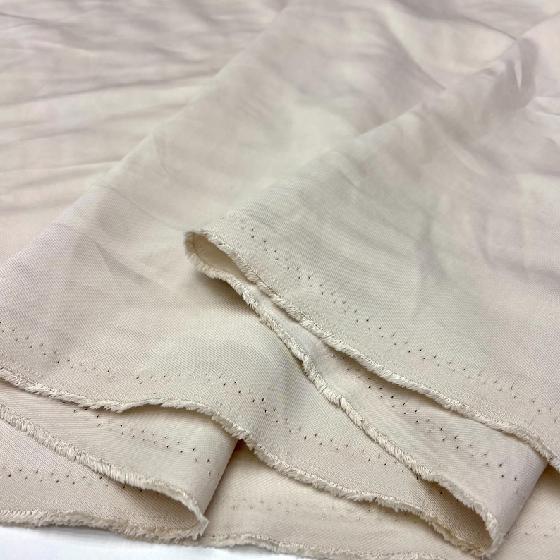 May include: A length of off-white, unbleached cotton fabric featuring a visible twill weave texture. The material is folded in soft, cascading layers, revealing a raw, frayed selvage edge with a series of small, evenly spaced pinhole marks.