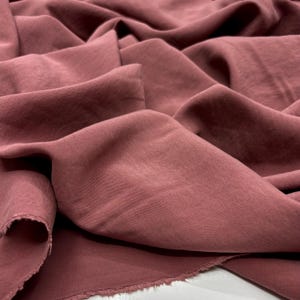 Puede incluir: Primer plano de una tela rosa empolvado con pliegues suaves y un acabado mate. La tela parece ser un material ligero, posiblemente seda o un textil similar y drapeable. El color es un tono cálido y apagado.