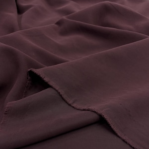 May include: A length of deep burgundy fabric with a smooth, matte texture and visible folds. The material features a raw, frayed edge along the cut side, showcasing the weave of the textile. The fabric is draped to create soft, flowing shadows.