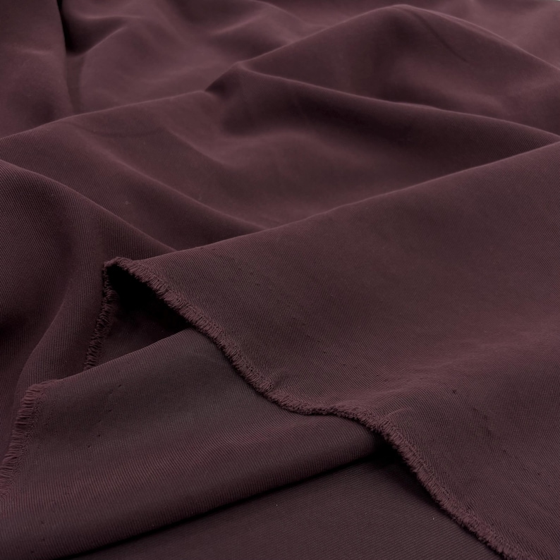 May include: A length of deep burgundy fabric with a smooth, matte texture and visible folds. The material features a raw, frayed edge along the cut side, showcasing the weave of the textile. The fabric is draped to create soft, flowing shadows.