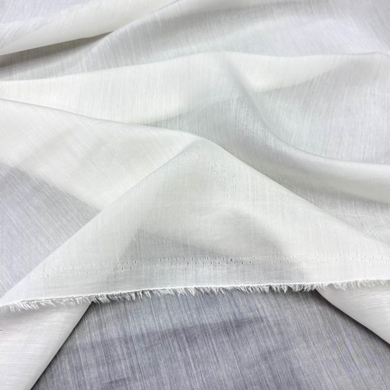 May include: Close-up of white fabric with a textured weave. The fabric is folded and draped, showing the texture and how it catches the light. The edges are unfinished, with a raw, fringed edge.