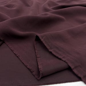 May include: A length of deep burgundy fabric with a soft, matte texture and visible folds. The material features a raw, frayed edge along the cut side, showcasing the weave of the textile. The fabric is draped to create gentle, flowing curves.