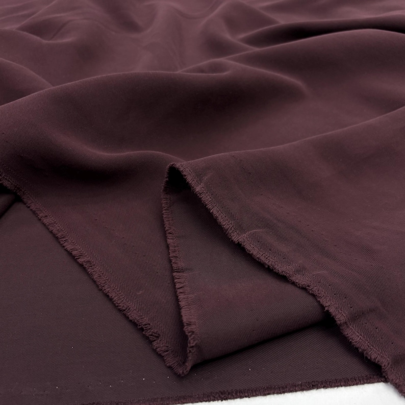 May include: A length of deep burgundy fabric with a soft, matte texture and visible folds. The material features a raw, frayed edge along the cut side, showcasing the weave of the textile. The fabric is draped to create gentle, flowing curves.