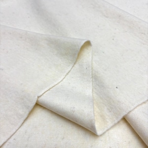 Puede incluir: Un trozo de tela de algodón crudo sin blanquear con una textura natural visible. El material presenta pequeñas motas de color marrón oscuro en todo el tejido, creando un aspecto moteado en la superficie plegada, suave y mate.