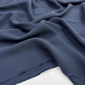 May include: A length of solid slate blue fabric with a soft, matte texture and a fluid drape. The material features visible folds and ripples across its surface, with a raw, unfinished edge showing a slightly frayed hem along the bottom border.