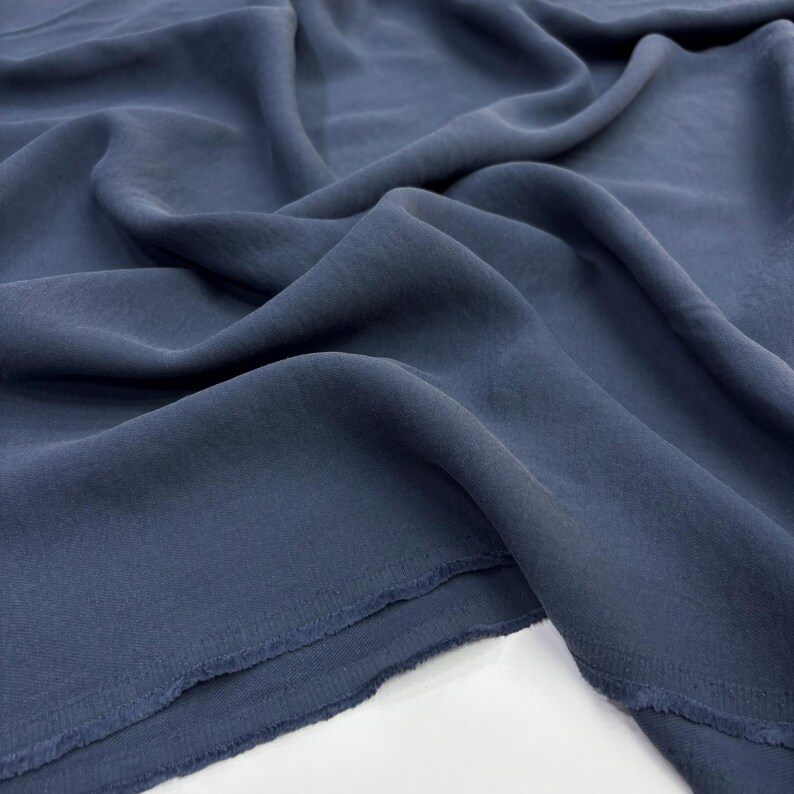 May include: A length of solid slate blue fabric with a soft, matte texture and a fluid drape. The material features visible folds and ripples across its surface, with a raw, unfinished edge showing a slightly frayed hem along the bottom border.