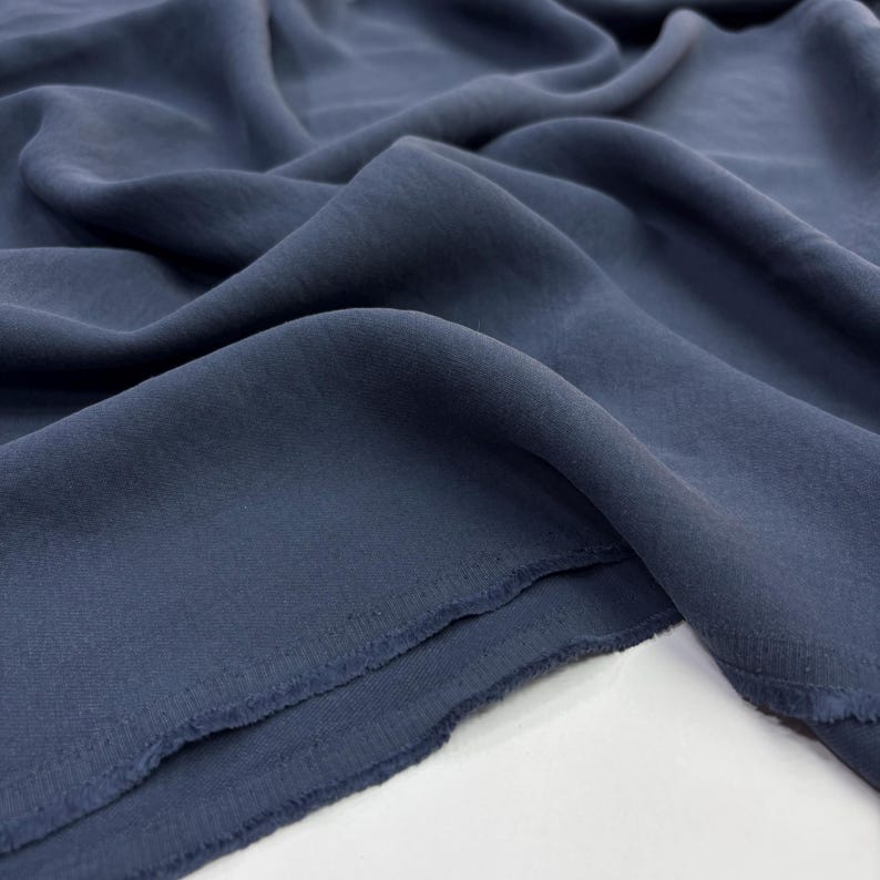 May include: A solid dark slate blue fabric with a matte finish and a soft, flowing drape. The material features visible folds and ripples across its surface, with a slightly frayed, raw edge visible along the bottom corner of the textile.