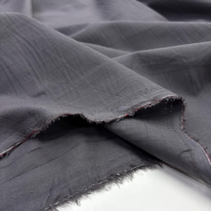 May include: Close-up of a gray fabric with a textured appearance. The fabric is draped, showing folds and a frayed edge with a hint of red. The material has small, evenly spaced perforations along one edge.